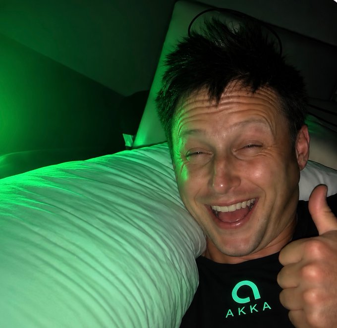 A cheerful man gives a thumbs-up while lying on a pillow bathed in green light, wearing a black "AKKA" t-shirt, enthusiastically supporting the AKKA prediction challenge tied to $HYPE's potential rise to $100.