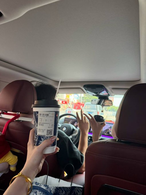 A hand wearing a gold bracelet holds a tall drink cup (order #10183) in the foreground of a car, while friends ahead high-five each other through the windshield in a celebratory weekend moment.