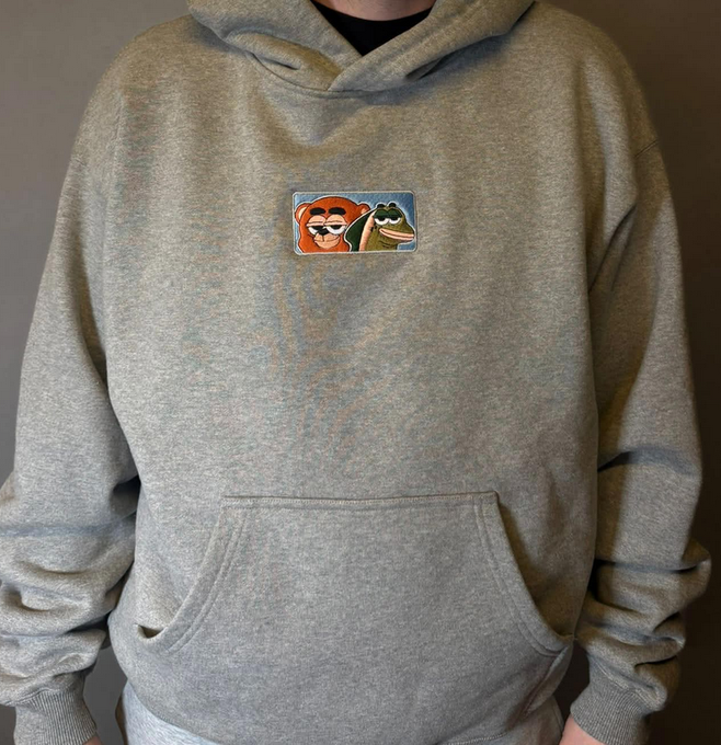 The image features a person wearing a gray hoodie that has an orange and green cartoon character design on the front, along with a small square patch of fabric containing two cartoon characters. The person appears to be standing against a plain white background. The text in the image reads "Who is interested in bears and salmon merch purchase with $BANDS + USDC?". This suggests that the hoodie might be related to merchandise for fans of bears, salmon, or other wildlife-related topics, possibly including items from the USDC (United States Department of Agriculture).