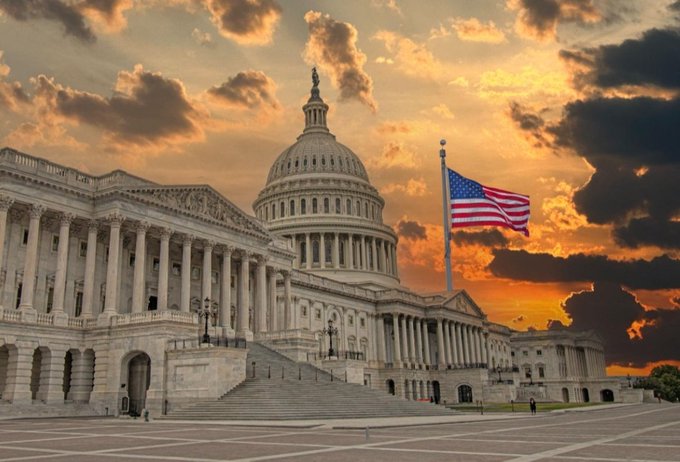 The image shows the U.S. Capitol building at sunset with an American flag waving prominently in the foreground, symbolizing the federal government's role in shaping policy. This visual ties directly to the post's theme of U.S. lawmakers pushing for clearer crypto regulations, as the Capitol represents the very institution where regulatory decisions affecting Bitcoin and the broader crypto market will be made. The dramatic, golden sky suggests a pivotal moment, reinforcing the idea that regulatory certainty could be the next major catalyst for Bitcoin's growth.