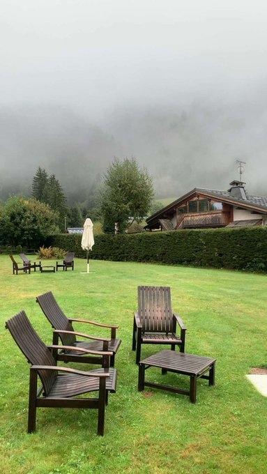 A misty mountain landscape with wooden outdoor lounge chairs, a patio table set, and a closed umbrella on a green lawn near a chalet, complementing the post's relaxed Saturday vibe from GM Legends on NomismaNetwork.