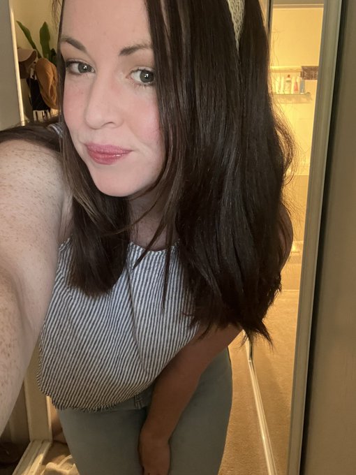 A woman with freckles and shoulder-length brown hair takes a mirror selfie wearing a striped sleeveless top and light green pants.