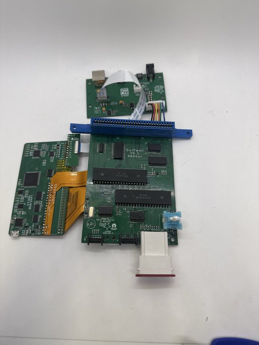 An electronics assembly featuring stacked green circuit boards connected by a white ribbon cable, an orange flexible flat cable, and a large D-sub connector rests on a white surface.