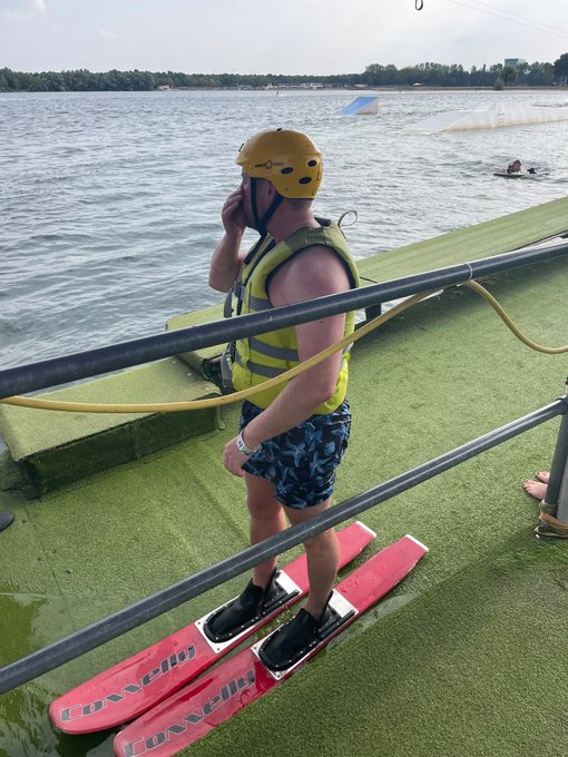 A person in a yellow helmet, life vest, and blue patterned shorts stands on red water skis on a floating dock at a lake with cable wakeboarding ramps visible in the background.
