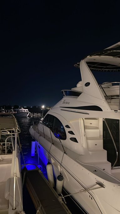 A nighttime photograph of a large white luxury yacht docked at a marina, illuminated by blue underwater lights, with "Sun Breeze" visible on its hull and other boats in the dark background.