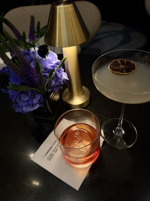 Two cocktails—one an amber Negroni-style drink and the other a clear cocktail garnished with a dried citrus slice—sit beside a small lamp and a vase of purple flowers at a table, with a napkin reading "coinbase" beneath them, capturing the upscale, celebratory setting of the Coinbase F1 Qualifying Watch Party where the meetup with the trading competition winner took place.