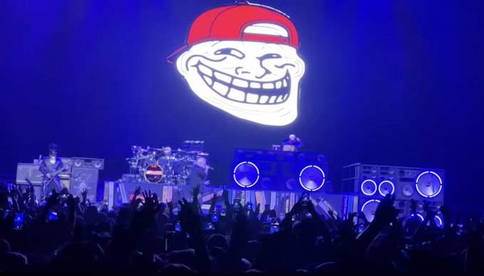 A live concert scene featuring the 'Trollface' meme graphic displayed on a large screen above a band performing on stage, with the audience's silhouettes and raised hands visible in the foreground.