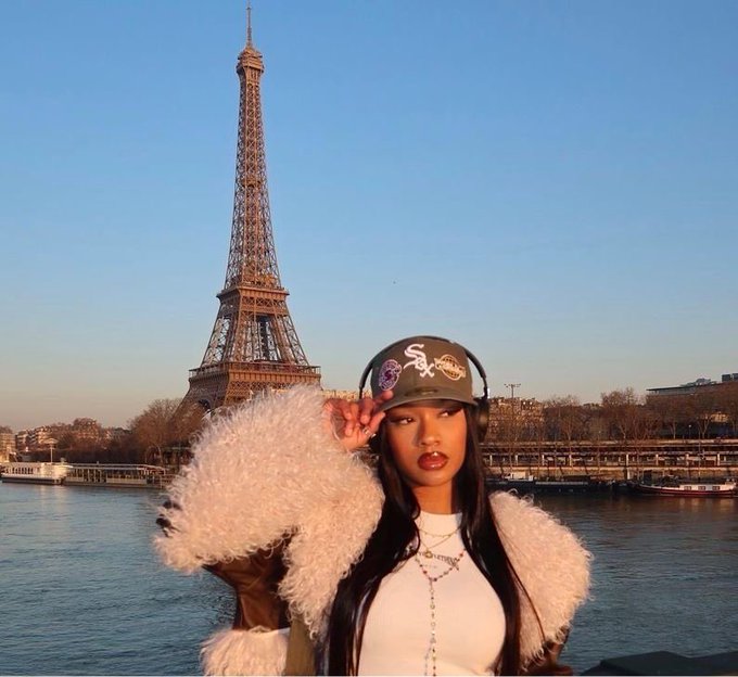 A woman wearing a white fuzzy jacket, White Sox cap, and headphones poses in front of the Eiffel Tower and the Seine River.