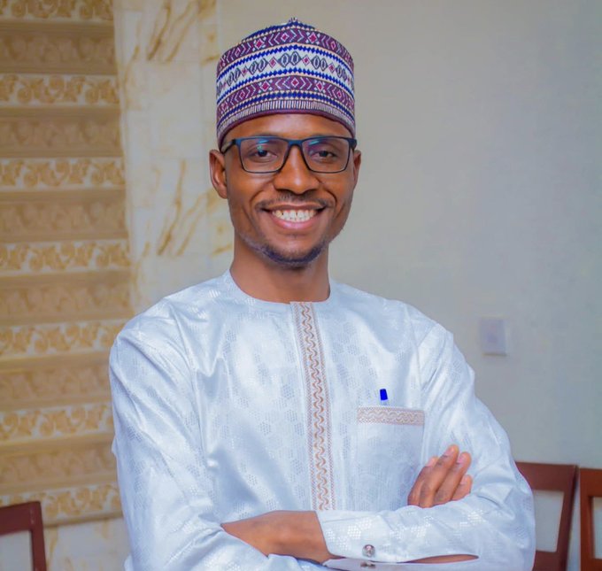 A smiling young man wearing glasses, a traditional embroidered cap (fula), and a white patterned kaftan with a pen in his chest pocket stands with arms crossed against a staircase background, posing confidently in what appears to be a professional or formal portrait associated with the "$SUNUSI" post.