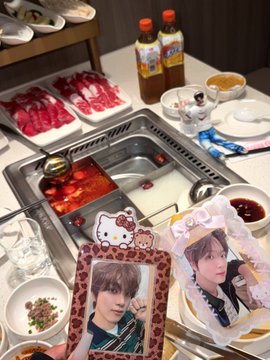 A HOTPOT dinner spread featuring a divided pot of spicy red broth and clear broth, surrounded by plates of sliced meat, condiments, and bottles of sauce, with a Hello Kitty placemat and two idol photo cards (one showing a member of HOLO) held up in the foreground. The image captures a fan's celebratory meal for HOLO's 1st Anniversary, combining the group-themed photo cards with a festive hotpot dinner.