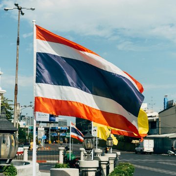The image shows a large Thai flag waving in the breeze against a blue sky. This relates to Thailand's SEC issuing rules allowing Bitcoin and Ethereum ETFs starting October 16th.