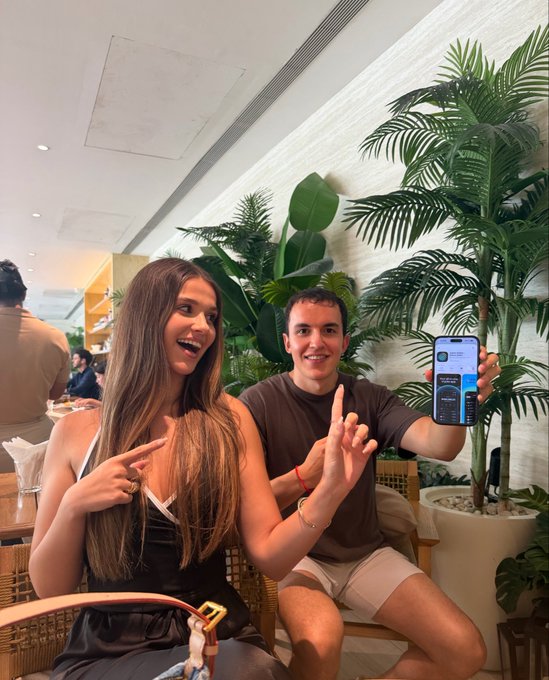 The image features a man and woman sitting next to each other with their hands on their laps while holding up a cell phone, possibly showing off something interesting or entertaining on the screen of the device. The background includes potted plants that add a touch of greenery to the scene. There is also another person visible in the background who appears to be engaged in conversation or observing something else.