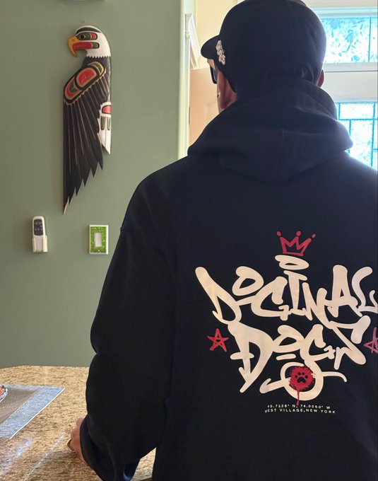 A person wearing a black hoodie with graffiti-style 'Decline' text on the back and a black cap, standing indoors near a wall decorated with a Northwest Coast Indigenous style carved eagle artwork.