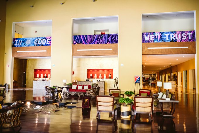 A spacious, modern hotel lobby with high ceilings features purple neon signs reading 'IN CODE' and 'WE TRUST' on the upper level, with seating areas, artwork, and warm lighting on the main floor.