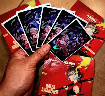 A hand holds up fanned-out Azuki NFT trading cards featuring anime character artwork, displayed above a red "Gates Awakened" Azuki TCG booster box in celebration of the trading card game release day.