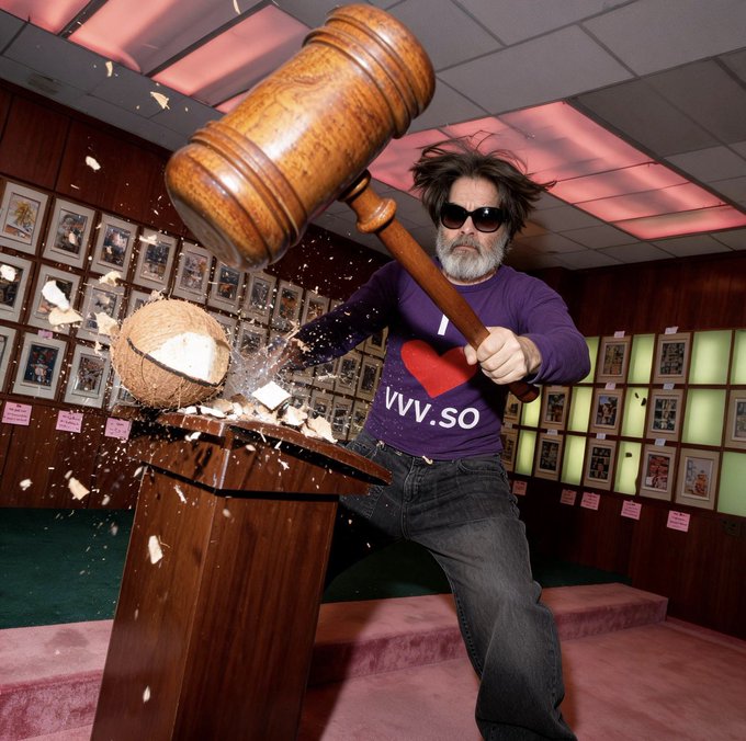 A man with a gray beard and sunglasses, wearing a purple "I [heart] VVV.so" shirt, swings a giant wooden mallet to smash a coconut on a podium, sending pieces flying.