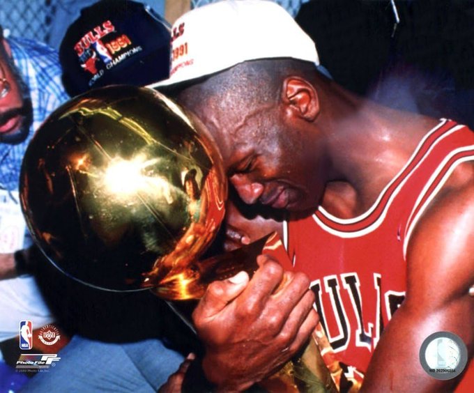 Michael Jordan in his red Chicago Bulls jersey kisses the 1991 NBA Championship trophy, with a 'Bulls 1991 World Champions' cap visible behind him.