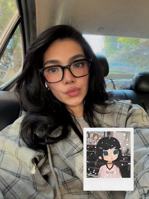A woman with long dark hair and black cat-eye glasses poses inside a vehicle, with a Polaroid-style photo of a cartoon avatar pinned to her plaid jacket.