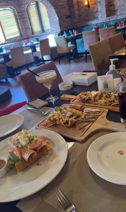 A restaurant table set with multiple plates of Mexican-style food including nachos and tacos, with a martini glass and other dishes visible.