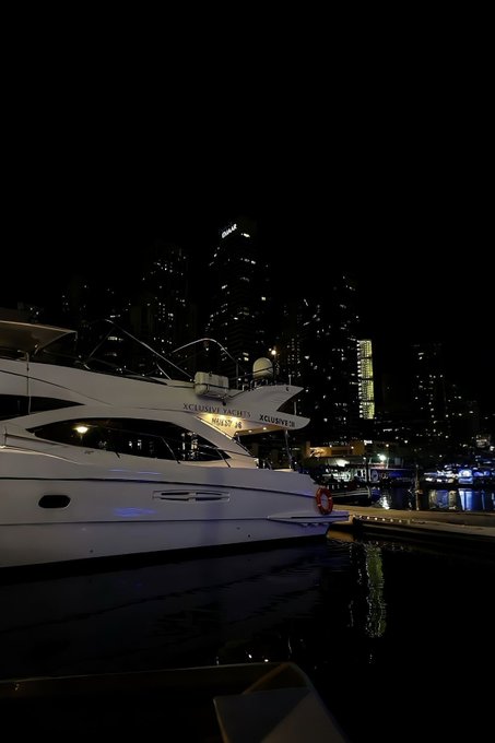 llor's yacht, a white boat with blue accents and the name "Lorien" on its side, is docked at a marina under a city skyline at night.
