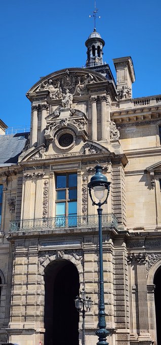 An ornate Renaissance-style stone building featuring intricate sculptures, a small bell tower, a circular window, and a classic street lamp in the foreground under a clear blue sky.