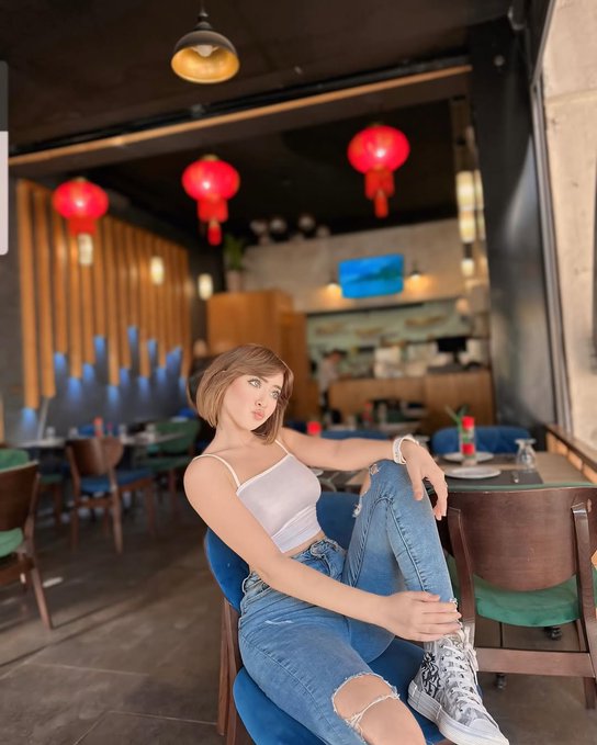 A young woman with short brown hair wearing a white tank top and ripped jeans sits casually at a wooden table inside a restaurant decorated with red Chinese lanterns.