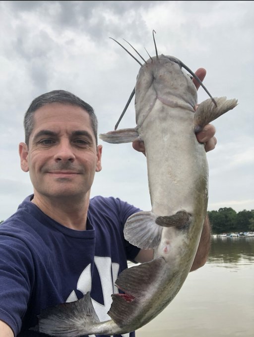 A selfie of a smiling man holding up a large catfish he caught, with a lake, trees, and cloudy sky in the background.