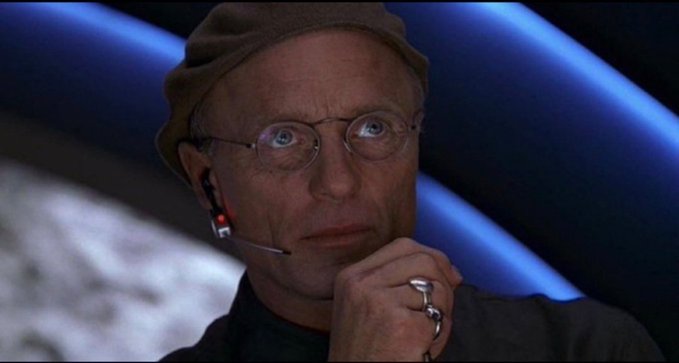 The image shows a thin, bald man with round glasses, a tan head covering, and a headset microphone, with his hand resting near his chin. The accompanying text references a deceptive financial manipulation tactic, mirroring the character's signature style of calculated deception and manipulation often portrayed in film.