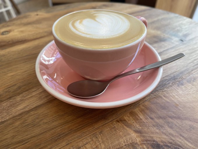 A pink ceramic cup filled with a latte featuring heart-shaped foam art sits on a matching saucer with a spoon on a wooden table.