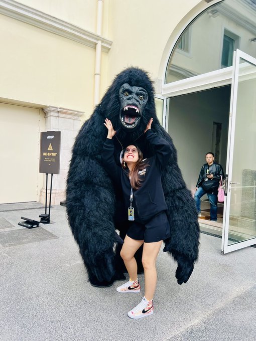 A woman poses playfully in front of a person in a King Kong-style gorilla costume, both striking dramatic poses outside a building. A sign reading "RE-ENTRY" is visible in the background, fitting the playful "Gm Apes" greeting perfect for crypto and NFT enthusiasts.