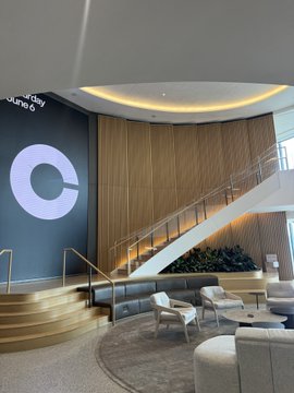 The image shows a spacious, modern lobby with a large wooden wall featuring a circular logo at its center. A white staircase leads up to this wall, providing easy access to the upper level of the building. In front of the staircase, there are two chairs and a couch arranged for seating. The floor is covered in tiles that reflect light throughout the space, creating an inviting atmosphere. On the right side of the image, there's a sign that reads "I think @coinbase won the nyc office competition".