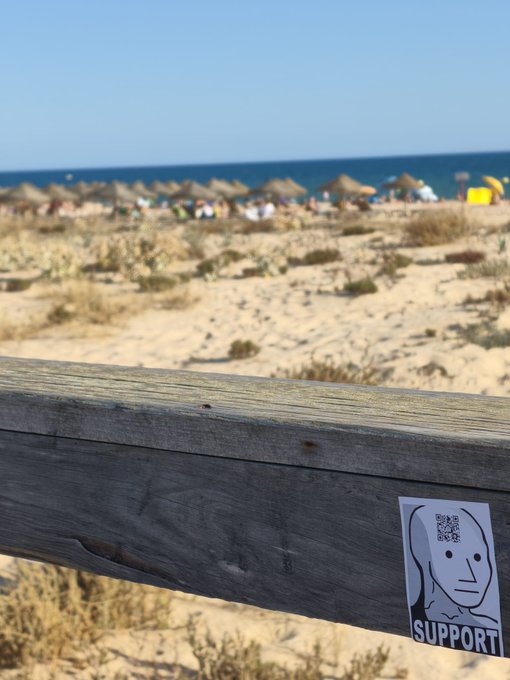 A wooden boardwalk railing in the foreground with a 'SUPPORT' sticker featuring a face illustration and QR code, overlooking a sandy beach with straw umbrellas and the ocean in the background.
