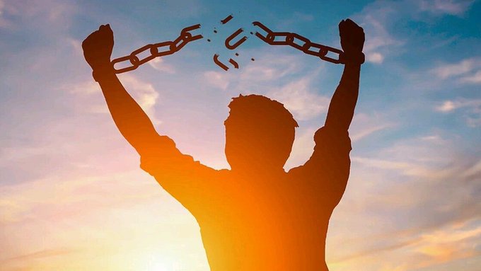A silhouette of a man in a red shirt breaking free from chains with his raised fists against a vibrant orange and blue sunset sky background.