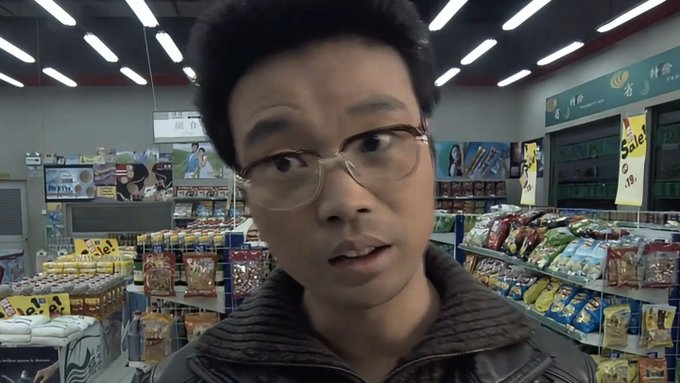 A man with glasses films himself inside a brightly lit convenience store, with shelves of snacks and sale signs visible behind him.
