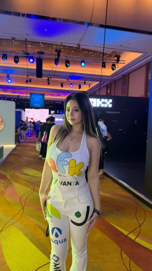 A young woman poses at what appears to be a crypto or tech convention, wearing a white tank top featuring 'OKX' branding and 'Vanta' sponsorship, paired with white pants displaying sponsor logos including Aqua.