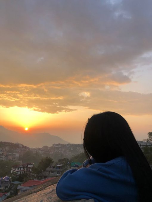The image features a woman with long hair, wearing a blue sweater, standing near a building or bridge overlooking a cityscape at sunrise. The sky has a beautiful orange hue as the sun rises over the horizon, creating a serene atmosphere. The woman is looking out towards the city and appears to be enjoying her time in this picturesque setting.