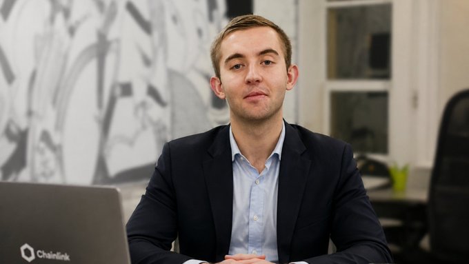 A young man in a dark suit jacket and light blue shirt sitting at a desk, with a laptop showing the 'Chainlink' logo in the foreground.