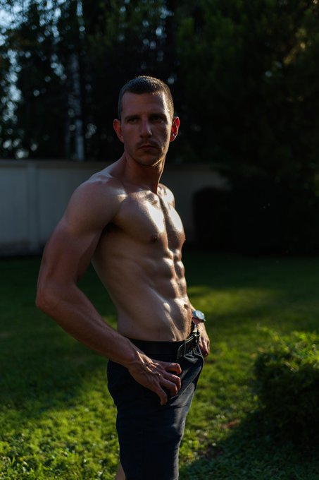 A shirtless, muscular man with short hair poses outdoors in a grassy area with trees and a fence in the background, wearing dark pants and a wristwatch.