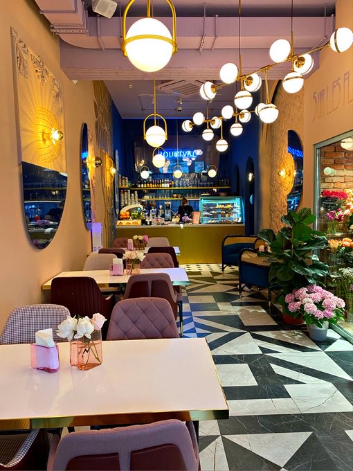 The image shows an empty restaurant with white tables, chairs, and pink flowers as decoration. The dining area has a black and white checkered flooring pattern. There are multiple tables set up throughout the space, each accompanied by two chairs. A potted plant is placed on one of the tables, adding to the ambiance. 

The restaurant appears to be empty at the moment, with no customers or staff present in the scene. The image also contains a caption that reads "Good Evening gQuip The best technology often works quietly in the background. That’s why infrastructure matters." This could possibly be an advertisement for Quip Network's services related to Web3 and blockchain technology.