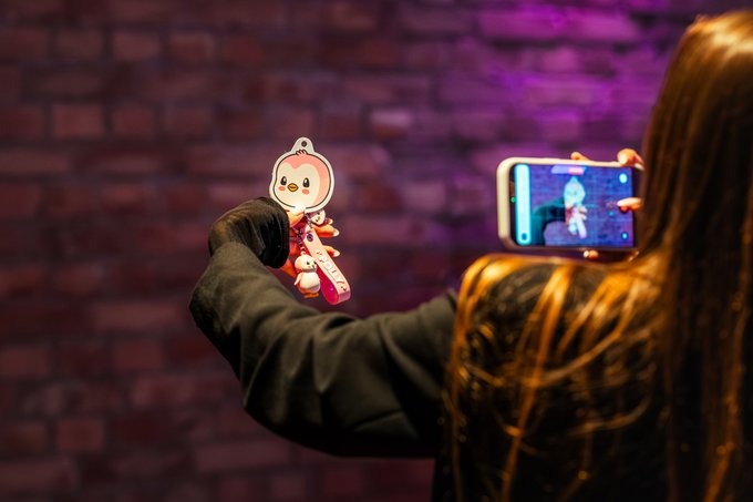 A person with long hair takes a photo of a cute Pudgy Penguins-themed phone charm/keychain against a pink-lit brick wall, with the smartphone screen displaying the figure being captured. The image ties directly to the post by showcasing a Pudgy Penguins collectible, tying in the partnership between Pudgy Penguins and GMC NFTs.