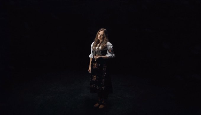 A young woman with long hair wearing a traditional dirndl dress with floral patterns stands alone in a dark, shadowy setting, looking upward with her hands clasped.