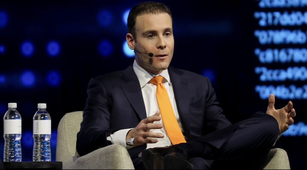 A man in a dark suit with an orange tie speaks while seated on a stage with a blue background displaying blurred white text and two water bottles on a table beside him.