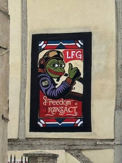 The image features a poster of a cartoon character, likely a frog or a bird, with a message that reads "Liberty, RANSACT", which translates to "Freedom, Ransom Act" in English. The poster is displayed on a building's exterior wall and is surrounded by various other posters and signs.