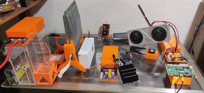 A workbench with various electronics including a stereo camera module, 3D printed parts in orange, circuit boards, and power supplies.