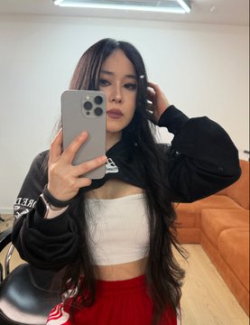 Mirror selfie of a young woman with long dark hair wearing a black jacket, white crop top, and red skirt, holding a smartphone.
