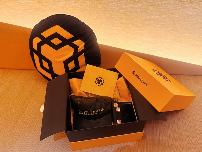 An unboxing of a BNB Chain merchandise box containing a black and yellow BNB logo pillow, a 'BUILDER' cap, a yellow card, and a smartphone on a wooden surface.