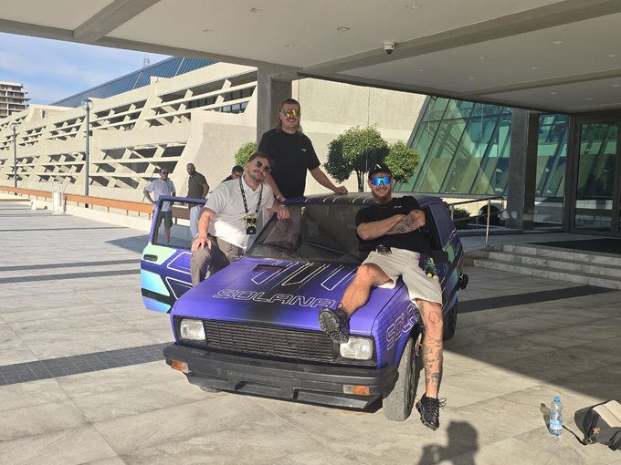 Three men pose on and around a small purple custom vehicle labeled "SCULPTOR," parked outside a modern building.