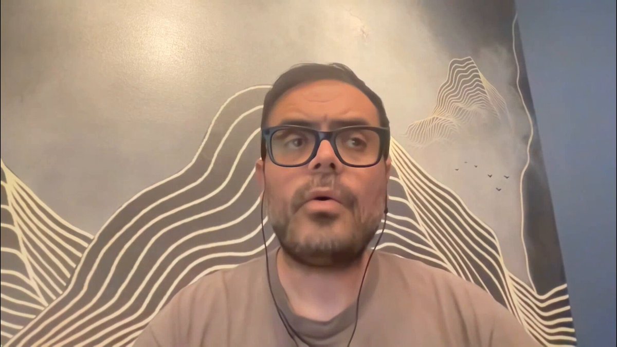 The image shows a man wearing glasses with a beard sitting at a desk or table. He appears to be looking directly into the camera while wearing headphones, suggesting that he might be participating in an online meeting or video call. The background features a painting of mountains on blue walls, which adds an artistic touch to the scene. The caption provided seems unrelated and does not provide any additional information about the image's content.