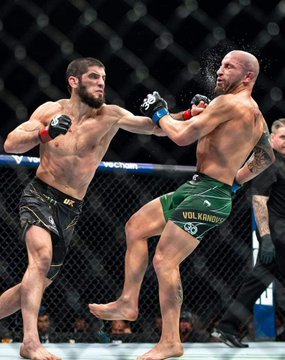 An action photograph from a UFC mixed martial arts fight showing a fighter in black shorts landing a punch on an opponent in green shorts inside the octagon.