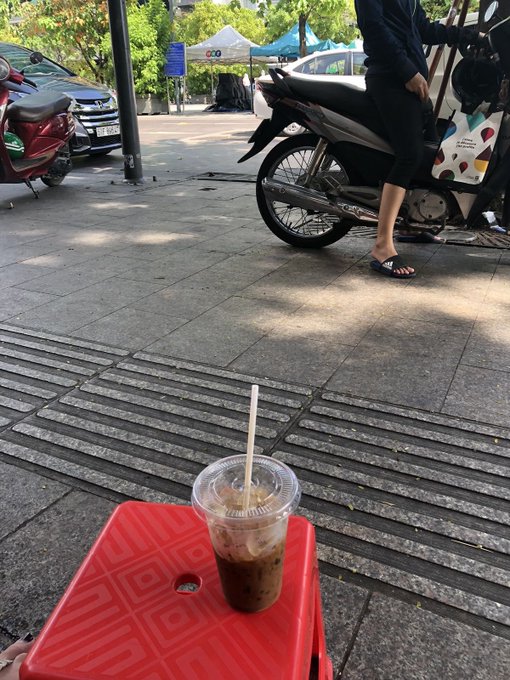 A cup of iced coffee with a straw rests on a red plastic stool in the foreground, while a person on a motorbike stands nearby on the sidewalk, with a blue car bearing license plate 76A-12345 and a white tent with 'coo' text visible in the background.